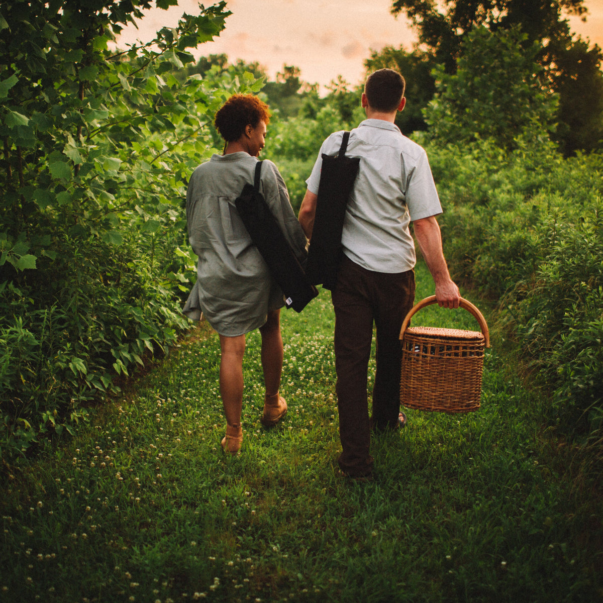 Two people walking through a forest with a picnic basket and carrying two Kermit Chairs folded up in their carrying cases.