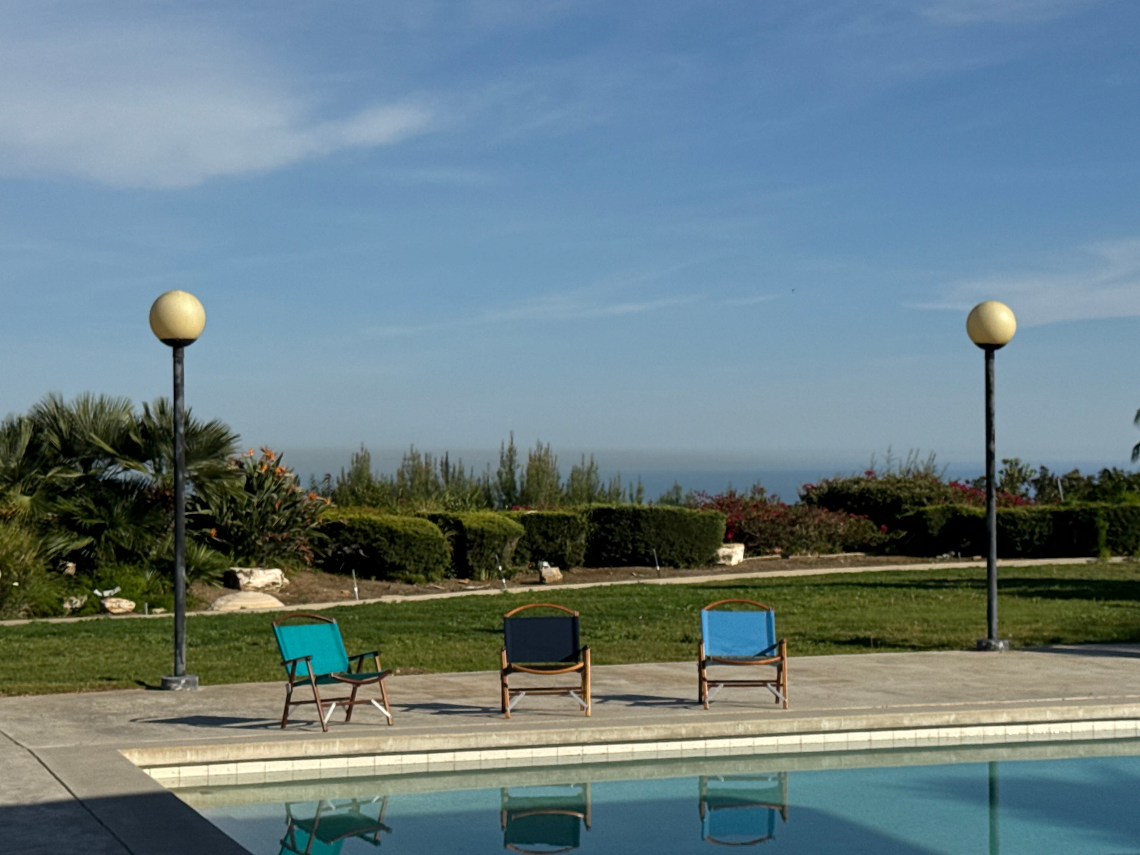 Three Classic Walnut folding Kermit Chairs by a pool designed by Richard Neutra, with a garden in the background.